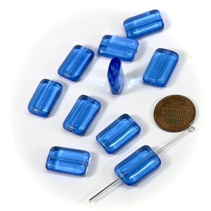 Könnte beinhalten: Zehn blaue Glasperlen, jede mit einer rechteckigen Form und einer leicht erhabenen Linie in der Mitte. Die Perlen sind auf einer weißen Oberfläche verstreut, mit einem Penny und einer Nadel als Größenvergleich.