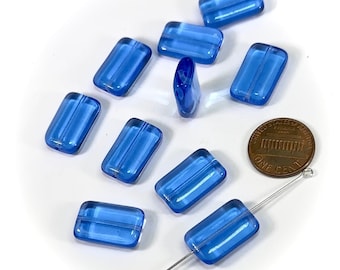 May include: A group of ten blue glass beads, each with a rectangular shape and a slightly raised line down the center. The beads are scattered on a white surface, with a penny and a needle for scale.