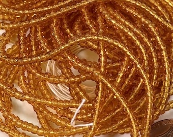 May include: Close-up of a pile of small, golden-colored glass beads. The beads are strung together, creating a tangled, textured appearance. The beads are a warm, amber color, and appear to be for jewelry making or crafting.