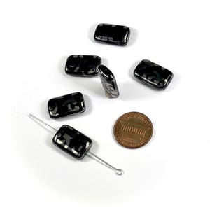 May include: Six black and silver rectangular glass beads with a patterned design. The beads are arranged on a white surface with a penny for scale.