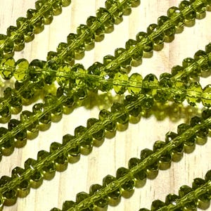 May include: Close-up of several strands of faceted, olive-green beads. The beads are strung together, creating a repeating pattern. The beads are translucent and reflect light, giving them a sparkling appearance. The background is a light-colored, wooden surface.