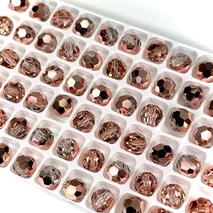 May include: Close-up of a white tray filled with rose-gold faceted beads. The beads are arranged in a grid pattern, each bead reflecting light. The beads have a metallic sheen, suggesting they are made of glass or crystal, and are ideal for jewelry making.