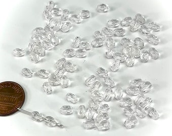 May include: A large quantity of clear glass beads, oval shaped with a faceted design. The beads are scattered on a white surface. A US penny is included for scale.