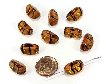 May include: A collection of amber-colored glass beads with black stripe designs. The beads are oval-shaped and vary slightly in size. A US one-cent coin is included for size comparison. A needle is inserted into one of the beads.