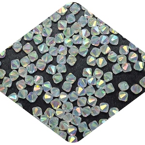 May include: A close-up of iridescent clear glass beads, also known as bicones, with a faceted design. The beads are scattered on a black surface.