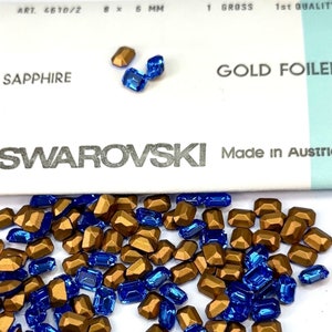 May include: A close-up of a pile of gold and sapphire blue Swarovski crystals. The crystals are in the shape of rectangular facets. The crystals are arranged in a random pattern. The crystals are on a white surface. The text "SWAROVSKI Made in Austria" is visible on the surface.