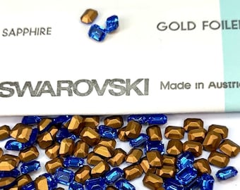 May include: A close-up of a pile of gold and sapphire blue Swarovski crystals. The crystals are in the shape of rectangular facets. The crystals are arranged in a random pattern. The crystals are on a white surface. The text "SWAROVSKI Made in Austria" is visible on the surface.