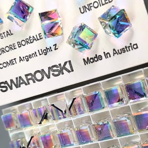 May include: A close-up of a white card with the text "SWAROVSKI" and "Made in Austria" printed on it. The card is displaying a variety of iridescent square crystals. The crystals are arranged in a grid pattern and are labeled "UNFOILED" and "Comet Argent Light Z."
