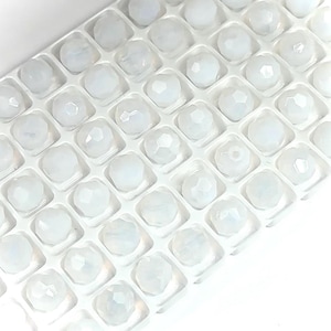 Op de afbeelding: Een close-up van een witte plastic container gevuld met kleine, witte, gefacetteerde glazen kralen. De kralen zijn gerangschikt in rijen en kolommen.