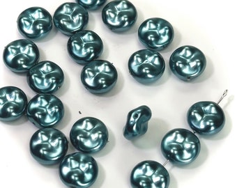 May include: A collection of teal-colored, round beads with a unique, textured surface. The beads have a lustrous, pearlescent finish and are scattered across a white background. Some beads are strung on a wire, suggesting they are for jewelry making.