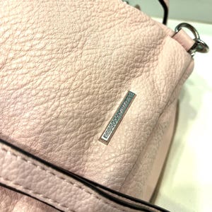 Op de afbeelding: Een lichtroze leren handtas met een getextureerde oppervlakte. De tas heeft een schouderriem en een zilverkleurig metalen label met de tekst "REBECCAMINKOFF". De tas heeft een zachte, pastelkleur.