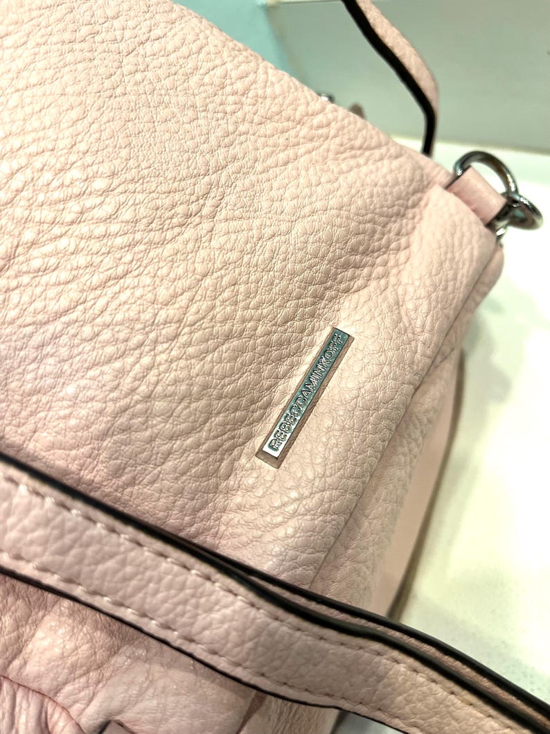 Op de afbeelding: Een lichtroze leren handtas met een getextureerde oppervlakte. De tas heeft een schouderriem en een zilverkleurig metalen label met de tekst "REBECCAMINKOFF". De tas heeft een zachte, pastelkleur.