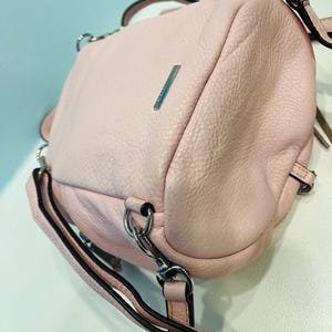 Op de afbeelding: Een lichtroze leren handtas met zilverkleurige hardware. De tas heeft een getextureerde oppervlakte, een handvat aan de bovenkant en een lange verstelbare riem. Een kleine zilveren balk is zichtbaar aan de voorkant. De tas kan over de schouder of als crossbody worden gedragen.