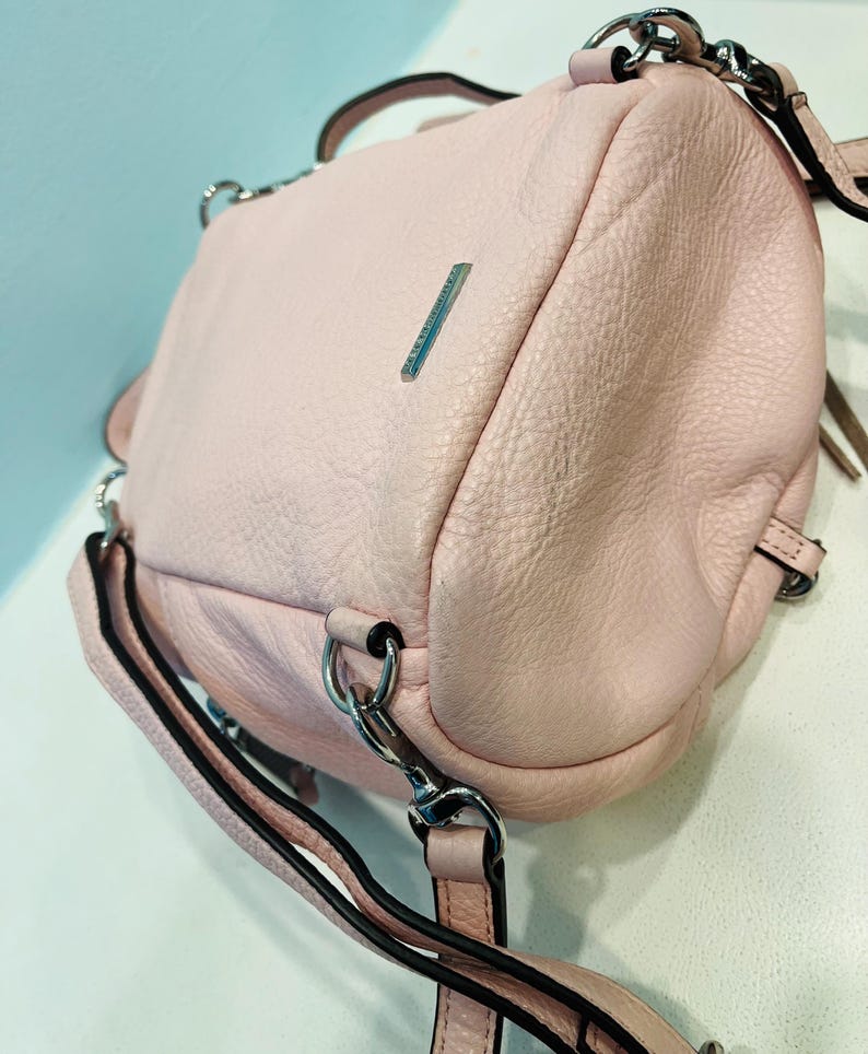 Op de afbeelding: Een lichtroze leren handtas met zilverkleurige hardware. De tas heeft een getextureerde oppervlakte, een handvat aan de bovenkant en een lange verstelbare riem. Een kleine zilveren balk is zichtbaar aan de voorkant. De tas kan over de schouder of als crossbody worden gedragen.