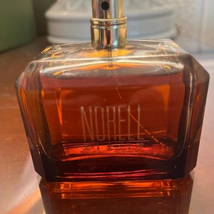 May include: A square-shaped perfume bottle with a gold-colored cap and the word "NORELL" printed on the front. The bottle is filled with a reddish-brown liquid and sits on a reflective surface.