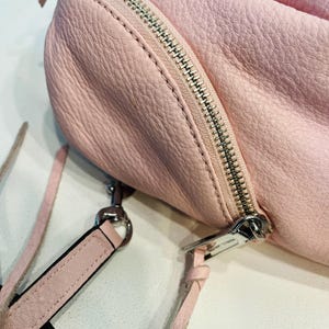 Op de afbeelding: Een lichtroze leren handtas met een zilveren rits en een afneembare schouderriem. De tas heeft een getextureerd oppervlak en een afgeronde vorm. De riem is ook lichtroze.