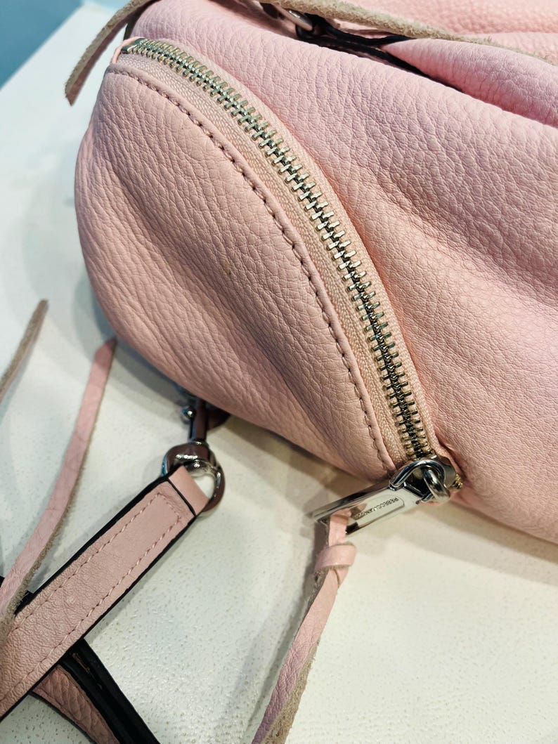 Op de afbeelding: Een lichtroze leren handtas met een zilveren rits en een afneembare schouderriem. De tas heeft een getextureerd oppervlak en een afgeronde vorm. De riem is ook lichtroze.