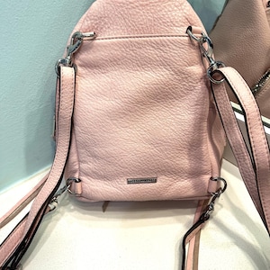 Op de afbeelding: Een lichtroze leren rugzak met zilverkleurige hardware. De kleine rugzak heeft verstelbare bandjes en een handvat aan de bovenkant. De achterkant van de tas heeft een klein vak en de merknaam is zichtbaar. De tas is gemaakt van getextureerd leer.