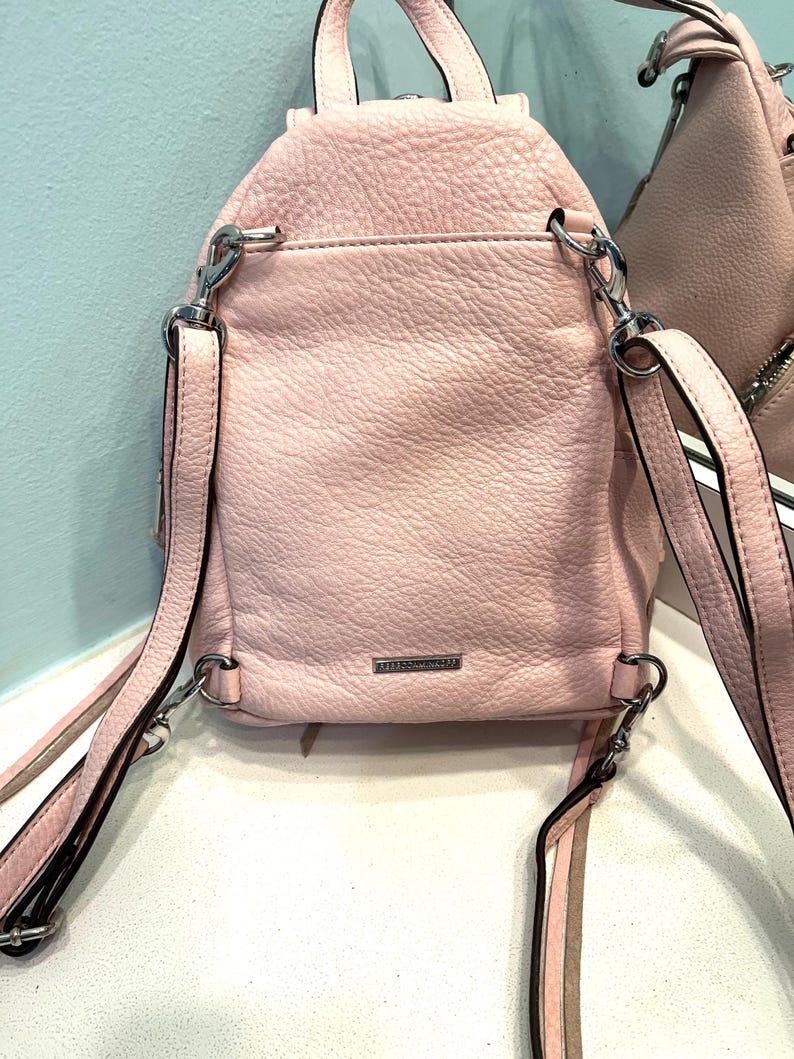 Op de afbeelding: Een lichtroze leren rugzak met zilverkleurige hardware. De kleine rugzak heeft verstelbare bandjes en een handvat aan de bovenkant. De achterkant van de tas heeft een klein vak en de merknaam is zichtbaar. De tas is gemaakt van getextureerd leer.