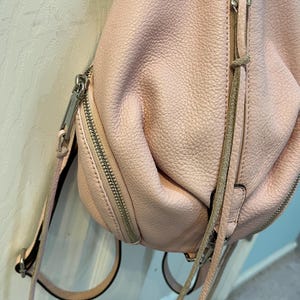 Op de afbeelding: Een lichtroze leren rugzak met zilveren ritsen en hardware. De tas heeft een getextureerd oppervlak en verstelbare bandjes. De ritsen hebben leren treklussen. De rugzak hangt aan een witte deur.