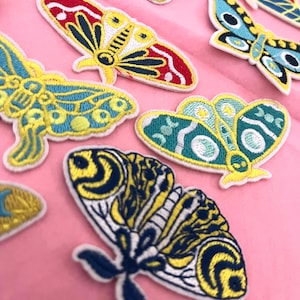 May include: Embroidered butterfly patches in various colors, including red, yellow, blue, and green, with intricate details. The patches are arranged on a pink surface, showcasing their vibrant designs and textures. These patches are ideal for adding a decorative touch to clothing or accessories.