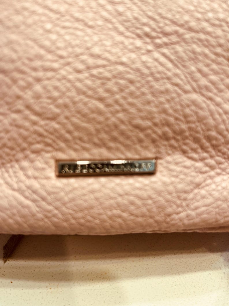 Op de afbeelding: Close-up van een lichtroze leren handtas. Het leer heeft een getextureerde, korrelige uitstraling. Een klein, rechthoekig zilverkleurig metalen label is aan de tas bevestigd.