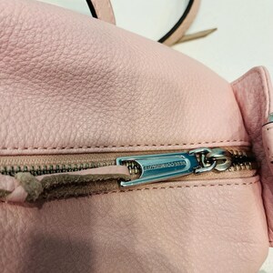 Op de afbeelding: Close-up van een lichtroze leren rugzak met een zilveren rits. De ritstrekker is gegraveerd met de woorden "REBECCA MINKOFF". De tas heeft een getextureerd oppervlak en een bijpassende roze riem.