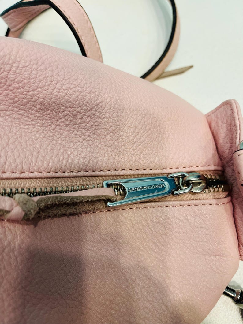 Op de afbeelding: Close-up van een lichtroze leren rugzak met een zilveren rits. De ritstrekker is gegraveerd met de woorden "REBECCA MINKOFF". De tas heeft een getextureerd oppervlak en een bijpassende roze riem.