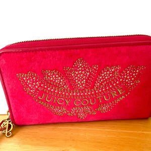May include: A bright pink wallet with a gold zipper and a gold-colored charm. The wallet features a large gold-colored crown design with the words "JUICY COUTURE" in gold lettering.