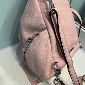 Op de afbeelding: Een lichtroze leren rugzak met zilverkleurige hardware. De tas heeft een ritssluiting, verstelbare bandjes en een handvat aan de bovenkant. Het leer heeft een getextureerde uitstraling. De tas is ontworpen om op de rug te worden gedragen.