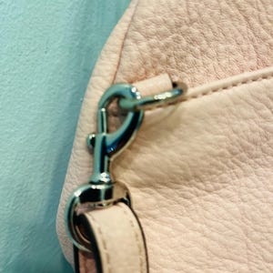 Op de afbeelding: Close-up van een lichtroze leren handtas met een zilverkleurige metalen sluiting en riembevestiging. Het leer heeft een getextureerde nerf en de tas lijkt nieuw te zijn.
