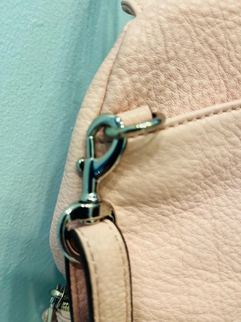 Op de afbeelding: Close-up van een lichtroze leren handtas met een zilverkleurige metalen sluiting en riembevestiging. Het leer heeft een getextureerde nerf en de tas lijkt nieuw te zijn.