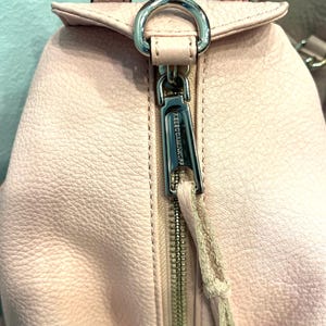 Op de afbeelding: Close-up van een lichtroze leren rugzak met een zilveren rits en hardware. De ritstrekker is een bijpassend leren koord. De merknaam is zichtbaar op de ritstrekker. Het leer heeft een getextureerde uitstraling.