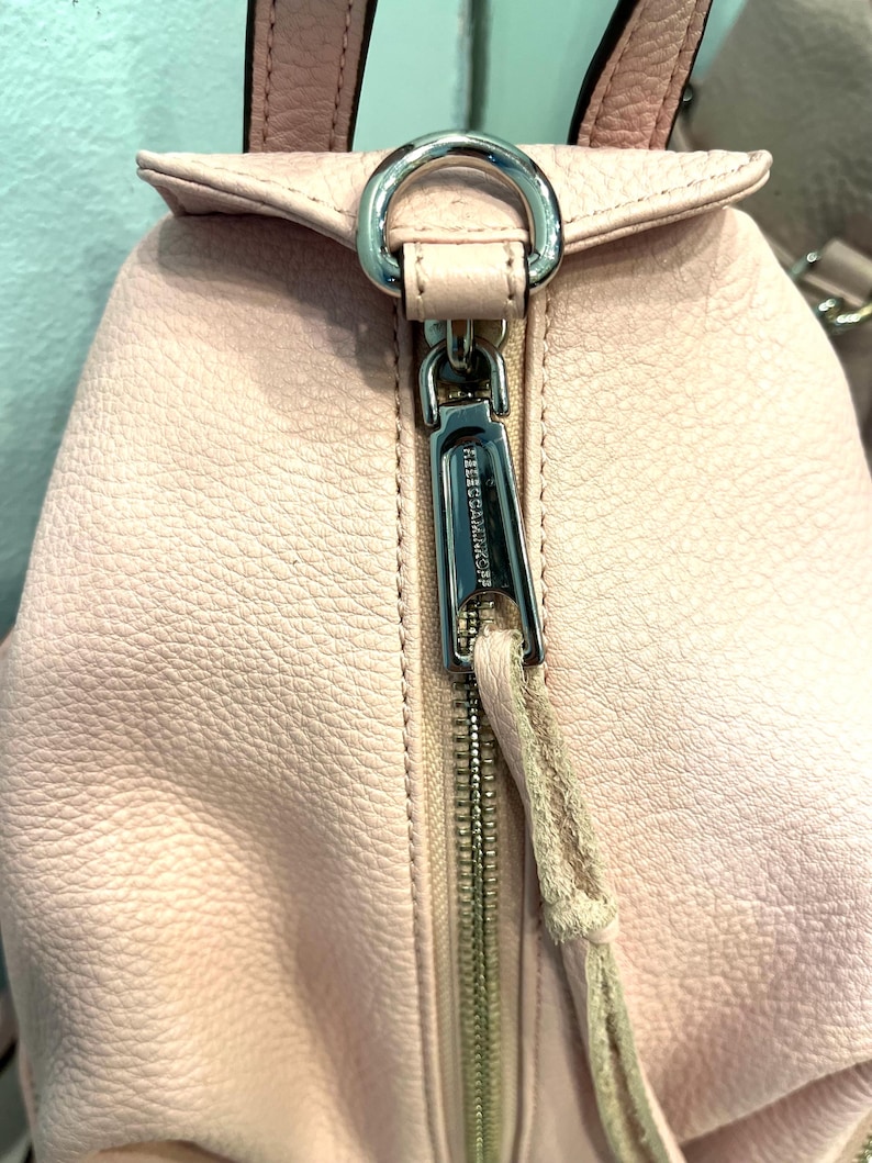 Op de afbeelding: Close-up van een lichtroze leren rugzak met een zilveren rits en hardware. De ritstrekker is een bijpassend leren koord. De merknaam is zichtbaar op de ritstrekker. Het leer heeft een getextureerde uitstraling.