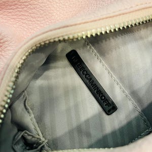 Op de afbeelding: Close-up van een lichtroze leren handtas. De binnenkant is grijs met een zwart label met de tekst "REBECCAMINKOFF". De tas heeft een zilveren rits en een zacht, getextureerd uiterlijk.