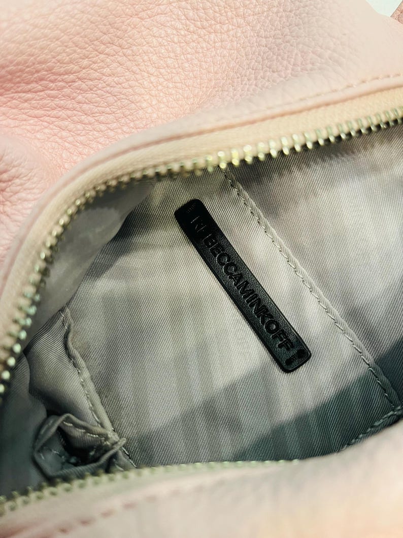 Op de afbeelding: Close-up van een lichtroze leren handtas. De binnenkant is grijs met een zwart label met de tekst "REBECCAMINKOFF". De tas heeft een zilveren rits en een zacht, getextureerd uiterlijk.