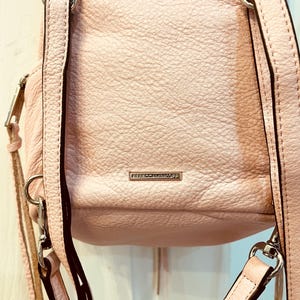 Op de afbeelding: Een lichtroze leren rugzak met zilverkleurige hardware. De tas heeft verstelbare bandjes en een klein rechthoekig metalen label met de merknaam. Het leer heeft een getextureerde uitstraling.