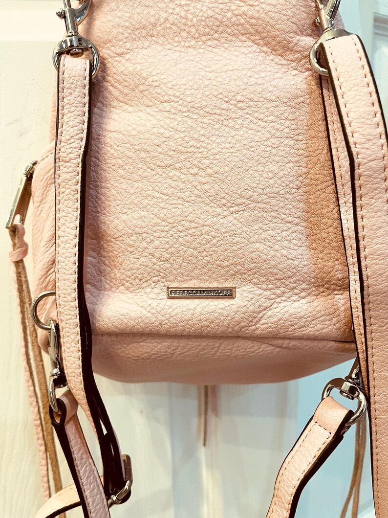 Op de afbeelding: Een lichtroze leren rugzak met zilverkleurige hardware. De tas heeft verstelbare bandjes en een klein rechthoekig metalen label met de merknaam. Het leer heeft een getextureerde uitstraling.