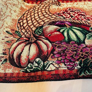 May include: A woven tapestry featuring a cornucopia overflowing with autumn harvest items. The design includes a woven basket, pumpkins, grapes, and other produce in shades of orange, red, green, and purple. The border is a deep red.