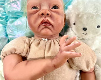 May include: A realistic baby doll with a neutral skin tone, wearing a beige onesie. The doll has detailed facial features and is lying next to a white stuffed sheep. The background features a light blue, textured fabric.