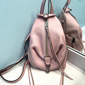 Op de afbeelding: Een roze leren rugzak met een handvat aan de bovenkant en verstelbare bandjes. De tas heeft zilverkleurige ritsen en hardware, met een klein ritsvakje aan de voorkant. De rugzak wordt weerspiegeld in een spiegel.