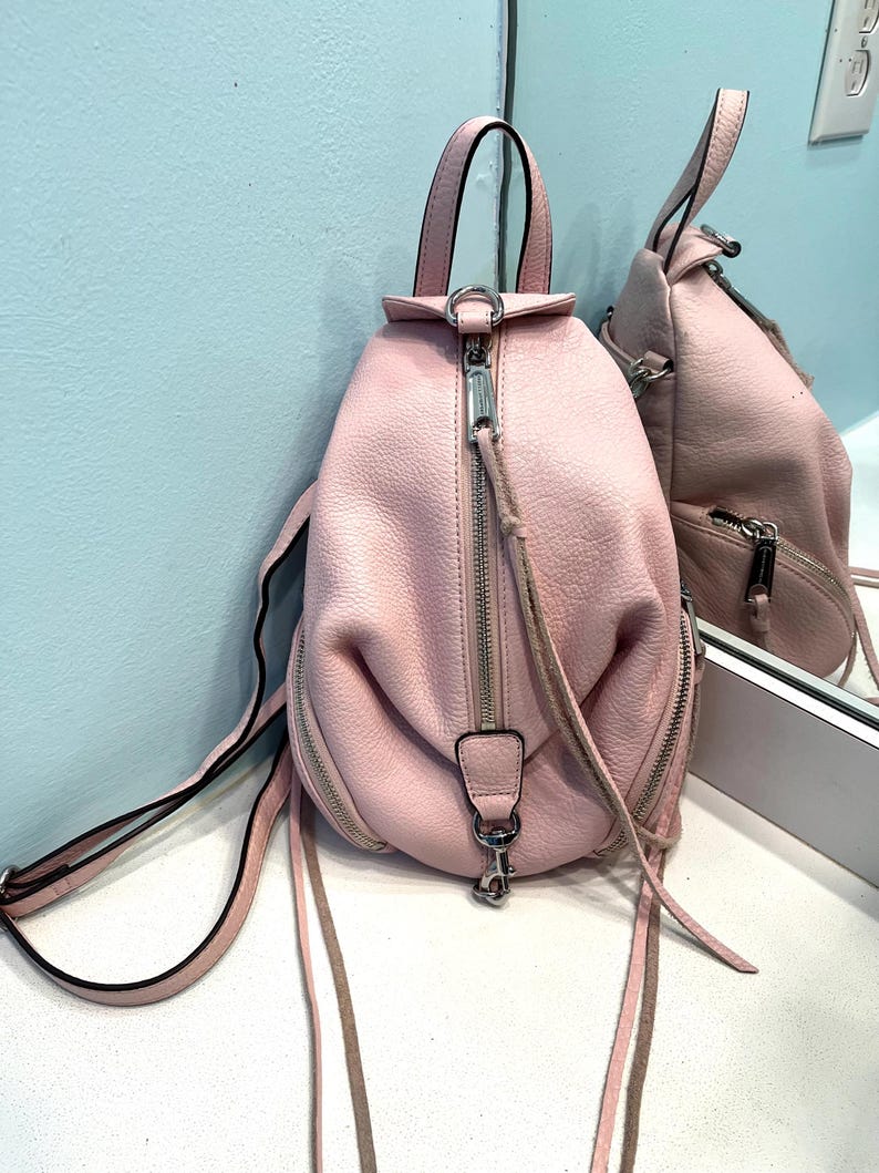 Op de afbeelding: Een roze leren rugzak met een handvat aan de bovenkant en verstelbare bandjes. De tas heeft zilverkleurige ritsen en hardware, met een klein ritsvakje aan de voorkant. De rugzak wordt weerspiegeld in een spiegel.