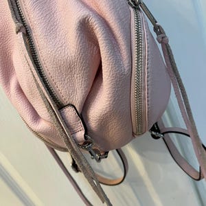 Op de afbeelding: Een lichtroze leren rugzak met zilveren ritsen en hardware. De tas heeft een getextureerd oppervlak en verstelbare bandjes. De ritsen zijn gesloten. De tas is zachtroze.