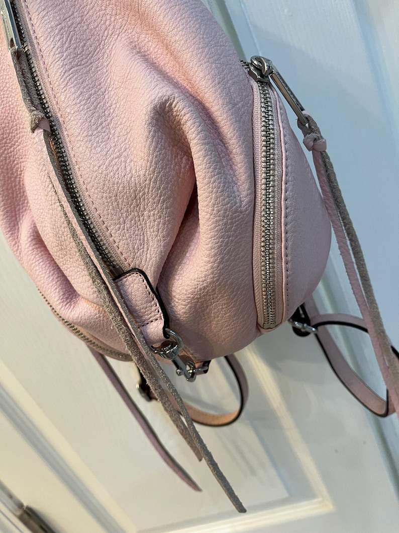 Op de afbeelding: Een lichtroze leren rugzak met zilveren ritsen en hardware. De tas heeft een getextureerd oppervlak en verstelbare bandjes. De ritsen zijn gesloten. De tas is zachtroze.