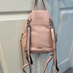 Op de afbeelding: Een lichtroze leren rugzak met zilverkleurige hardware, hangend aan een deurklink. De rugzak heeft verstelbare bandjes en een handvat aan de bovenkant. Het leer heeft een getextureerde uitstraling.