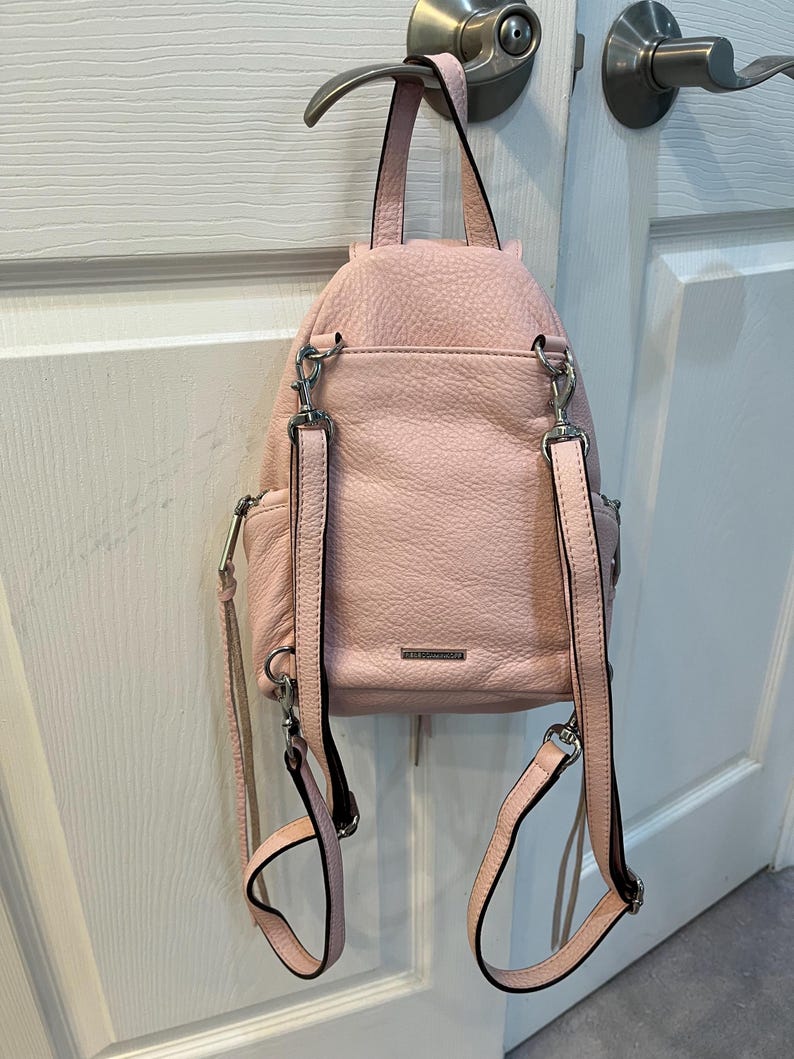 Op de afbeelding: Een lichtroze leren rugzak met zilverkleurige hardware, hangend aan een deurklink. De rugzak heeft verstelbare bandjes en een handvat aan de bovenkant. Het leer heeft een getextureerde uitstraling.