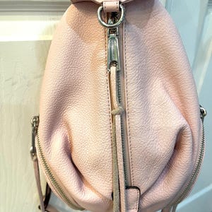 Op de afbeelding: Een lichtroze leren rugzak met zilverkleurige hardware. De kleine rugzak heeft een handvat aan de bovenkant, een ritssluiting aan de voorkant en twee ritssluitingen aan de zijkanten. De tas heeft een getextureerd oppervlak en een kleine kwast.