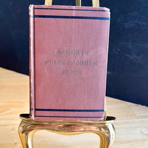 May include: A vintage book titled "Roberts Rules of Order Revised" with a dusty rose cover and dark blue stripes. The book is displayed on a small, ornate gold-colored stand, with a dark background.