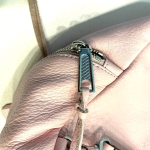 Op de afbeelding: Close-up van een lichtroze leren tas met een zilveren rits. De ritssluiting is zilverkleurig en heeft een textuurontwerp. Een dunne, lichtbruine riem is zichtbaar, samen met zilveren hardware.