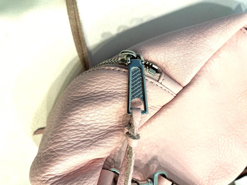 Op de afbeelding: Close-up van een lichtroze leren tas met een zilveren rits. De ritssluiting is zilverkleurig en heeft een textuurontwerp. Een dunne, lichtbruine riem is zichtbaar, samen met zilveren hardware.